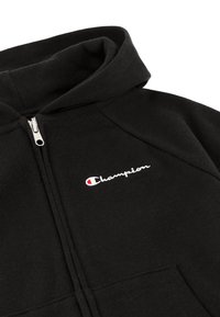 Sudadera negra con cremalleras hecha de tela suave, que presenta una capucha acanalada, una cremallera en la parte frontal y un logo bordado en blanco de "Champion" con un acento rojo.