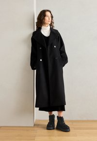 Manteau noir croisé avec grands revers, poches latérales et boutons argentés ; porté sur un col roulé. Chaussures : bottines noires à semelles épaisses.