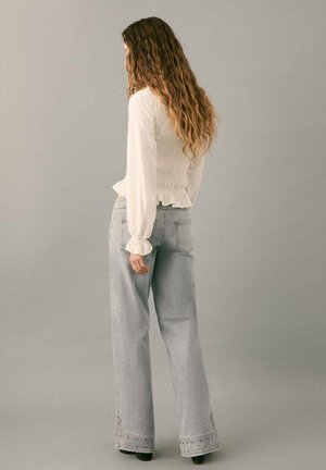 Donna con capelli lunghi e ondulati indossa una camicetta bianca a maniche lunghe e jeans di denim chiaro a vita alta e gamba larga, in piedi davanti a uno sfondo grigio semplice.