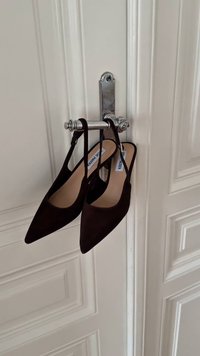 Brune semsket lær-slingbacks med spisse tær, hengende på en krom dørkrok. Har et minimalistisk design og glatt tekstur.
