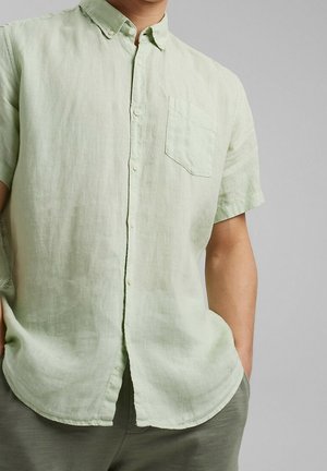 Mężczyzna ubrany w jasnobeżową koszulę na krótki rękaw z kieszenią na piersi, z rękami w kieszeniach, stojący na tle jednolitego szarego tła.