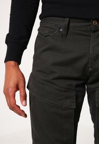 Cargo byxor i mörkgrön tyg, med sidofickor, bältesömmar, knäppning med knapp och en slät yta. Handen vilar på midjebandet.