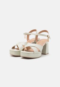 Tacones de plataforma verde claro con correas cruzadas y un tacón bloque. Presentan un acabado texturizado y un pequeño detalle de cadena.