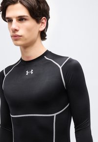 Νεαρός άνδρας που φοράει μαύρο μακρυμάνικο συμπιεστικό μπλουζάκι με λευκές ραφές και γκρίζο λογότυπο Under Armour στο στήθος.