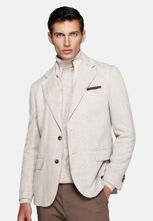 Young man wearing a light beige checkered blazer over a cream cable-knit turtleneck sweater with brown pants, hand in pocket.
