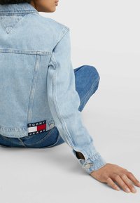 Světle modrá džínsová bunda volného střihu se stříbrnými knoflíkovými detaily a nášivkou "TOMMY JEANS" na zadní straně. Dlouhé rukávy s knoflíkovými manžetami.