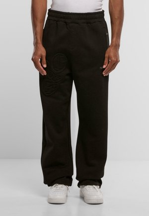 Black sweatpants made of soft fabric, featuring an elastic waistband and embossed logo details on the left leg. Styled with white sneakers.