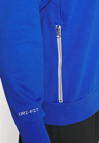 Manche de veste de sport bleue avec texte blanc "DRI-FIT" et une poche zippée verticale argentée au-dessus de la ceinture, portée avec un pantalon noir.