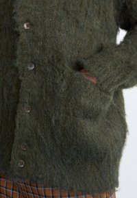 Ελιά πράσινη, φuzzy κάρντιγκαν φτιαγμένο από υφαντό μάλλινο υλικό με κουμπιά μπροστά και πλαϊνές τσέπες. Φορεμένο πάνω από καρό καφέ και μπλε παντελόνι.