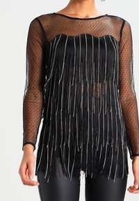 Haut noir avec un motif en tulle à pois, orné de longues franges et d'accents argentés. Encolure ronde et manches longues transparentes.