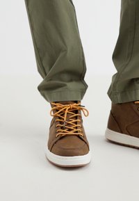 Zapatillas altas marrones con cordones naranjas, parte superior de cuero sintético texturizado y suela de goma blanca. Pantalones verde olivo parcialmente visibles.