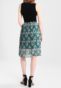 Top nero senza maniche abbinato a una gonna plissettata teal con motivi floreali bianchi. La modella indossa tacchi neri con cinturino alla caviglia.