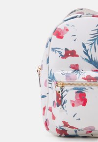 Kwiatowy plecak na białym tle, z wzorami różowych i niebieskich kwiatów, złotymi okuciami oraz przednią kieszenią zapinaną na zamek.