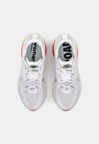 Baskets de sport blanches avec une tige en mesh, détails texturés gris, talon rouge et éléments réfléchissants. Semelle extérieure crantée pour une meilleure adhérence.