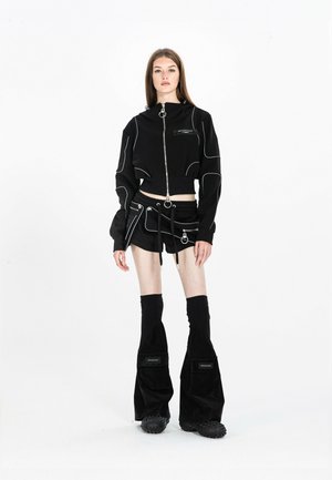 Mujer que lleva una chaqueta negra corta con cremallera, pantalones cortos negros con bolsa adjunta y pantalones negros acampanados con bolsillos, de pie sobre un fondo blanco.