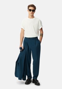 Witte t-shirt met opgerolde mouwen, donker tealkleurige geplooide broek, zwarte zonnebril en stevige zwarte schoenen, met een bijpassende jas in de hand.