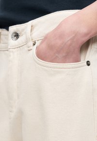 Hand in die Vordertasche einer cremefarbenen Jeans aus Denim gesteckt, mit sichtbaren Metallknopf- und Nieten-Details.