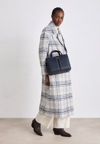 Musta-valkoinen ruudullinen pitkä takki, joka on yhdistetty tummansiniseen käsilaukkuun, jossa on teksturoitu pinta ja vaaleanpunaisia yksityiskohtia. Ruskeat korkokengät viimeistelevät asun.