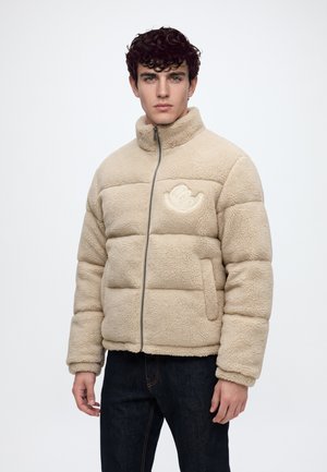 Jeune homme aux cheveux bouclés et foncés porte une veste polaire matelassée beige avec fermeture éclair et logo sur la poitrine, associée à un pantalon foncé sur fond uni.