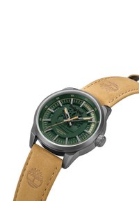Reloj con esfera verde, caja de acero inoxidable plateado, correa de cuero marrón y diseño texturizado. Presenta manecillas luminiscentes y grabado del logotipo.