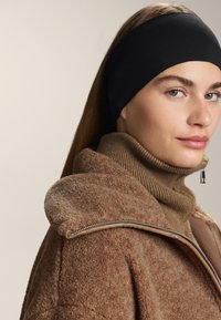 Černý látkový pásek na hlavu, který se nosí s béžovým pleteným rolákem a hnědou fleecovou bundou s detaily stříbrného zipu.