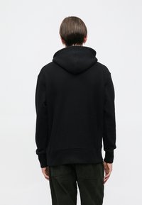 Čierna mikina s kapucňou vyrobená z mäkkej látky. Má rebrované manžety a spodný lem, voľný strih a bez viditeľných log. Zobrazený je zadný pohľad.