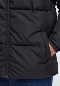 Jachetă neagră tip puffer, cu închidere prin fermoar, textură matlasată orizontal și buzunare laterale. Material neted cu finisaj mat.