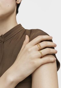 Złote pierścionki z misternymi wzorami, w tym łańcuchami i akcentami, są noszone na palcach w zestawieniu z neutralną brązową bluzką z kołnierzykiem.