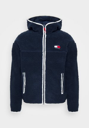 Person iført en navy fleecejakke, hvide sneakers, mørkeblå jeans og en lille navy crossbody taske, stående udendørs i et urbant område.