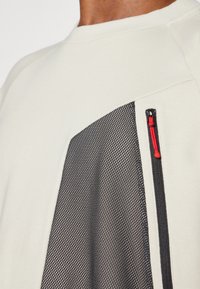 Light gray sweatshirt featuring a black mesh panel with a zip pocket, accented by a red pull tab and ribbed neckline and cuffs.