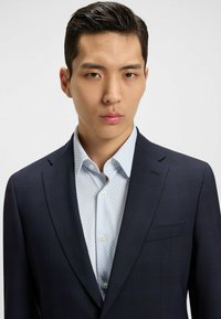 Marinblå kavaj med texturerad tyg, en bröstficka, bärs över en ljusblå skjorta med subtilt prickmönster och knäppt krage.