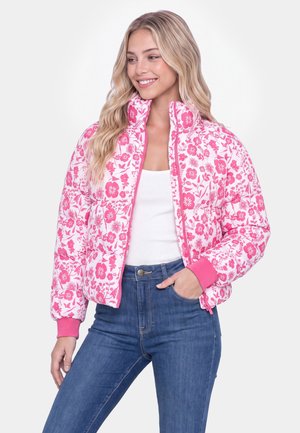 Frau trägt eine weiß-rosa geblümte Steppjacke, ein weißes Tanktop und blaue Jeans, lächelt und schaut zur Seite mit der Hand in der Tasche.