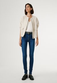 Chaqueta bomber beige con cremallera frontal y dos bolsillos, llevada sobre una camiseta blanca y jeans ajustados azules, combinada con zapatos negros con cordones.