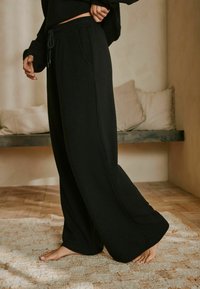 Schwarze weit geschnittene Hose aus strukturiertem Stoff, mit einem verstellbaren Bund und seitlichen Taschen. Das Modell steht barfuß auf einem strukturierten Teppich.