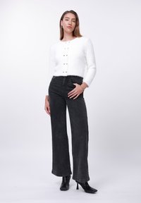 Λευκή fluffy ζακέτα με κουμπιά, σε συνδυασμό με ψηλόμεσα μαύρα denim παντελόνια με φαρδιά πόδια και μαύρες μυτερές μποτάκια αστραγάλου. Απλός σχεδιασμός.