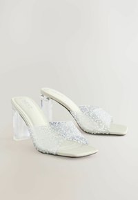Mules à talons hauts transparents avec un talon bloc transparent, ornés de perles blanches et bleues sur une sangle texturée.
