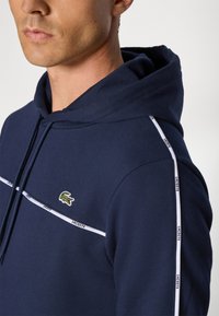 Sötétkék kapucnis póló pamut keverékből, kenguru zsebbel. Fehér csíkokkal és egy kis Lacoste krokodil logóval a mellrészen.