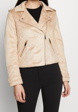 Woman wearing a beige quilted faux suede biker jacket with zippered pockets and black pants, standing against a plain background.