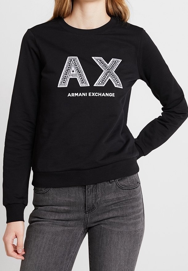 Sweat-shirt noir à manches longues avec les logos "AX" et "Armani Exchange", porté par une personne avec une main sur la hanche et un jean gris.