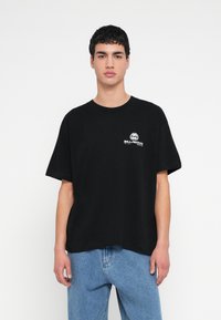 Czarny bawełniany t-shirt o luźnym kroju, z krótkimi rękawami i białym okrągłym logo na lewej stronie klatki piersiowej. Jednolite tło.