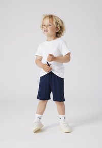 Bambino che indossa una maglietta bianca di cotone e pantaloni corti blu scuro, con calze bianche e scarpe chiare, in piedi con un'espressione giocosa.