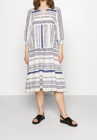 Vit klänning med blå geometriska mönster, trekvartslånga ärmar, V-ringning och volangkjol. Matchas med svarta sandaler.