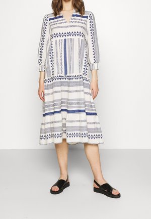 Valkoinen mekko sinisillä geometrisilla kuvioilla, kolmesta neljänneksestä hihat, V-pääntie ja kerrostettu hame. Yhdistetty mustien sandaalien kanssa.