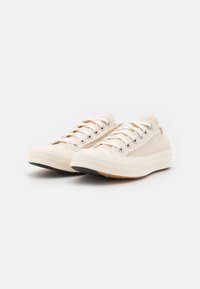 Baskets en toile beige avec lacets blancs, semelle en caoutchouc et flanc texturé, présentant un bout rond et des éléments de design minimalistes.