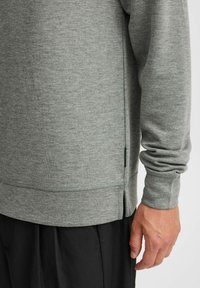 Grå, texturerad sweatshirt med långa ärmar och sidofickor. Har en rundad nederkant och ribbade ärmslut, tillsammans med svarta byxor.
