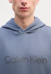 Pelēks kapuce ar teksturētu logo "Calvin Klein" tumšākā pelēkā. Iespējota klasiskā kapuce un maiga, gluda auduma. Nav rakstu vai akcentu.