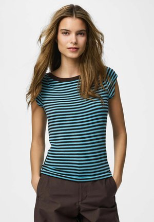 Ung kvinde med langt bølget hår iført en blå og sort stribet kortærmet top og mørke bukser, stående med hænderne i lommerne.