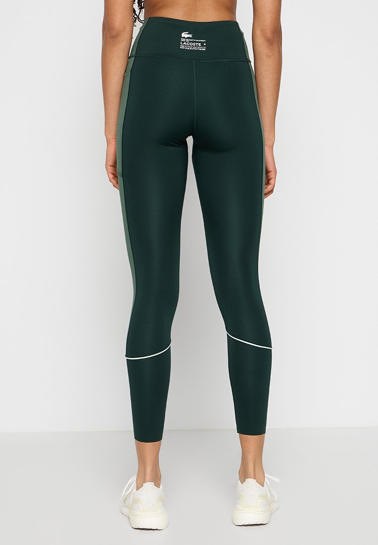 Leggings taille haute en vert foncé, fabriqués en tissu extensible. Présentent des coutures d'accentuation blanches et un logo subtil sur la taille.