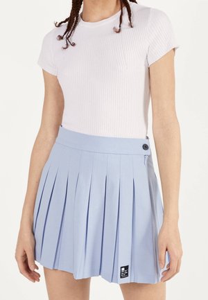 Lichtblauwe geplooide rok met een gladde textuur, voorzien van een zijknop en een klein zwart label. Gecombineerd met een aansluitend wit ribbelshirt.