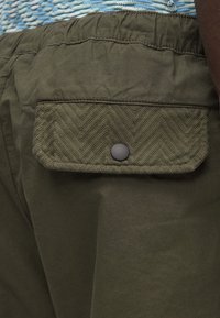 Pantalones cortos cargo de color verde oliva con un bolsillo con parche texturizado y cierre de botón a presión. La tela presenta una textura suave y ligera.